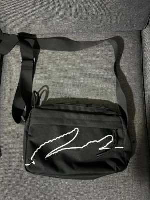 Svart axelväska från Lacoste - Snygg svart axelväska från Lacoste med justerbar rem och dragkedja. Väskan har ett vitt krokodilmotiv på framsidan och är tillverkad i slitstarkt tygmaterial. Perfekt storlek för det viktigaste och enkel att bära över axeln.