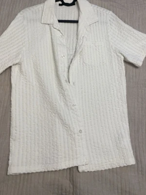 Vit kortärmad skjorta med struktur - Säljer en vit skjorta med korta ärmar och strukturerat tyg. Skjortan har klassisk krage, bröstficka och knäppning framtill. Perfekt för dig som gillar en clean och enkel stil med lite extra textur.