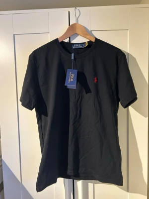 Svart t-shirt från Polo Ralph Lauren - Klassisk svart t-shirt från Polo Ralph Lauren med rund hals och korta ärmar. T-shirten har den ikoniska röda broderade loggan på bröstet och är tillverkad i mjuk bomull. Perfekt basplagg med stilren design. Helt ny priset kan diskuteras 