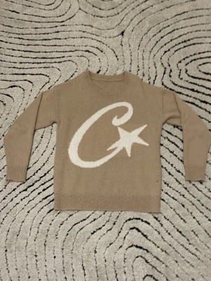 Beige stickad tröja med stjärnmotiv croteiz  - Beige stickad tröja med stor vit C och stjärna på framsidan. Tröjan har rund halsringning, långa ärmar och ribbstickade muddar vid ärmslut och nederkant. Perfekt för en avslappnad och trendig look.