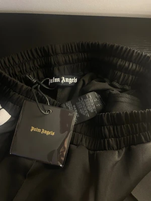 Svarta shorts från Palm Angels - Svarta shorts från Palm Angels med elastisk midja och vit sidoremsa. Klassisk streetwear-look med logotyp på insidan av midjan. Perfekta för en avslappnad och trendig stil.