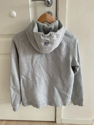 Ljusgrå hoodie  - Snygg ljusgrå hoodie med huva och ribbade muddar vid ärmsluten. Tillverkad i mjukt bomullsmaterial och har en liten broderad logga på huvan. Perfekt för en avslappnad och stilren look.