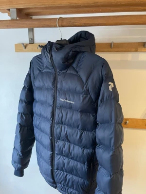 Mörkblå dunjacka från Peak Performance. Pris går att diskutera  - Snygg mörkblå dunjacka från Peak Performance med huva och dragkedja framtill. Jackan har diskret logga på bröstet och ärmen, samt quiltad design för extra värme. Perfekt för kalla vinterdagar och riktigt skön passform.