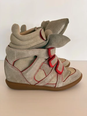 Beige sneakerskängor med kardborre - Unika beige sneakerskängor i mocka med coola röda detaljer och flera kardborreband. Skorna har en chunky sula i gummi och perforerade partier vid tårna för extra stil. Perfekta för dig som vill sticka ut med en sportig och trendig look.