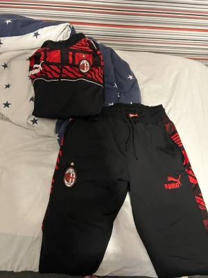 AC Milan träningsset från Puma - Säljer ett svart och rött AC Milan träningsset från Puma. Setet består av en ziptröja och byxor med klubbmärke och Puma-logga. Snygga röda detaljer och mönster längs ärmar och ben. Perfekt för fotbollsträning eller chill. 