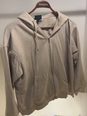 Beige zip hoodie från Divided - En stilren beige hoodie med dragkedja från Divided. Hoodien har en klassisk huva med dragsko, två fickor framtill och ribbade muddar vid ärmslut och nederkant. Tillverkad i mjukt bomullsmaterial som känns skönt mot huden. Perfekt för en avslappnad look.
