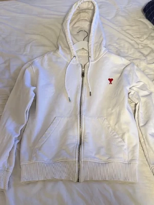 Vit zip hoodie från Ami Paris - Säljer en vit hoodie från Ami Paris med dragkedja framtill och huva med snören. På bröstet finns det klassiska röda Ami-hjärtat med bokstaven A. Hoodien har ribbade muddar och fickor på sidorna. Perfekt för en clean och stilren look.