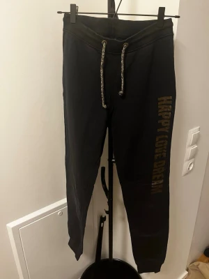 Svarta mjukisbyxor med tryck - Svarta mjukisbyxor med snörning i midjan och texten 'HAPPY LOVE DREAM' i guld på ena benet. Byxorna har muddar vid bensluten och är tillverkade i ett mjukt material som passar perfekt för en avslappnad stil.