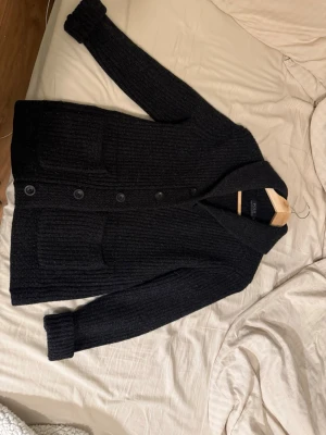Mörk Grå Cardigan tröja från Polo Ralph Lauren - Mysig mörk grå cardigan från Polo Ralph Lauren i grovstickad ullmix. Koftan har stora knappar framtill, två rejäla fickor och en bred sjalkrage. Perfekt till en mysig look, går även att klä upp, storlek S
