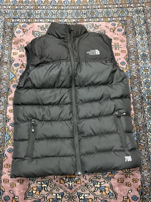 Svart dunväst från The North Face - Snygg svart dunväst från The North Face med hög krage och två dragkedjeförsedda fickor. Västen har en quiltad design och 700-fill power, perfekt för kalla dagar. Logga på bröstet och diskret 700-märkning nedtill.