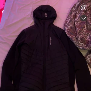  Sail Racing jacka  - Svart hybridjacka från Sail Racing med huva och dragkedja. Jackan har quiltad front och släta ärmar, samt diskret logga på bröstet. Materialmix med polyester och nylon ger en sportig vibe. Perfekt för dig som gillar stilren och funktionell design.