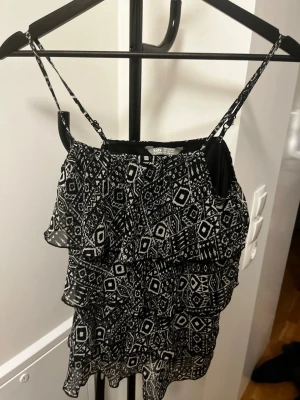Svartvit mönstrad linnetopp med volanger - Säljer en svartvit linnetopp med tunna axelband och grafiskt mönster. Toppen har flera lager volanger som ger en luftig och trendig look. Perfekt för dig som gillar detaljer och vill ha något som sticker ut.