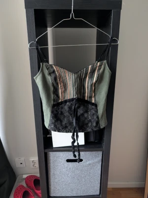 Grön och randig bustier med spets - Trendig bustier med smala svarta axelband, grön tygsida och randigt mönster framtill i beige, svart och rosa. Nedre delen framtill har svart spetsdetalj och snörning. Dragkedja bak. Perfekt för att skapa en edgy look.