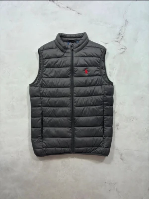 Ralph lauren väst - Snygg grå dunväst från Polo Ralph Lauren med quiltad design och dragkedja framtill. Västen har en klassisk ståkrage och den ikoniska röda logotypen broderad på bröstet. Perfekt för lager-på-lager och streetstyle. Ända defekten är att size tagen är av därför billig