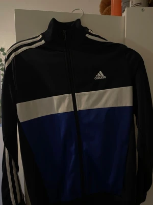 Svart och blå Adidas tränings zip hoodie  - Snygg träningsjacka från Adidas i svart och blått med vita detaljer och klassiska ränder på ärmarna. Dragkedja framtill och hög krage. Perfekt för sport eller chill. Adidas-logga på bröstet och mjukt material.