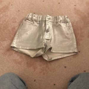 Silvriga metallicshorts med fickor - Snygga silvriga shorts i metallic-look med hög midja och klassisk femficksdesign. Shortsen har bälteshällor och knappstängning framtill. Perfekta för dig som vill sticka ut med en glittrig vibe.