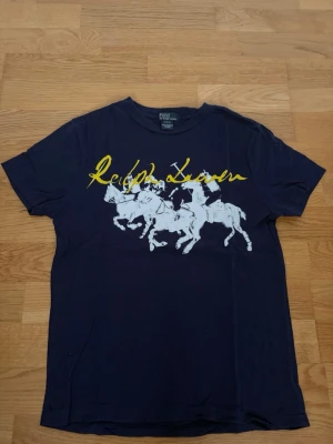 Mörkblå Ralph Lauren t-shirt - Vintage finns inte att köpa ny någonstans! Säljer en snygg mörkblå t-shirt från Ralph Lauren med stort tryck av polospelare i vitt och gul logga framtill. Klassisk passform och rund halsringning. Perfekt för dig som gillar stilrena plagg. Storleken är M i barnstorlek 14-16 år och motsvarar ungefär S i herr.
