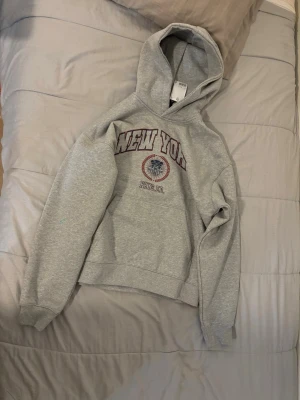 Grå hoodie med New York-tryck - Snygg grå hoodie med huva och trycket 'NEW YORK' i vinrött och vitt på bröstet. Under trycket finns även en liten emblem och texten 'BRKLYN, USA'. Hoodien har klassisk passform och ribbade muddar vid ärmslut och nederkant. Perfekt för en avslappnad streetstyle-look.