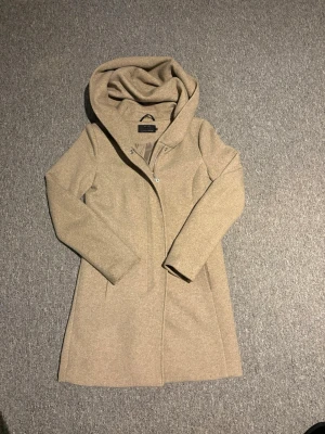 Beige kappa med huva från ONLY - Stilren beige kappa från ONLY med stor, mysig huva och dold knäppning framtill. Kappan har en rak passform och är tillverkad i ett mjukt tyg som ger en clean och modern look. Perfekt för dig som vill ha en enkel men snygg jacka till hösten eller våren.