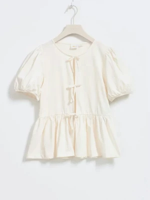 Krämvit blus med rosett och puffärm - Söt krämvit blus med korta puffärmar och två rosetter framtill. Blusen har en volang nedtill och rund halsringning. Tillverkad i ett lätt och luftigt tyg, perfekt för en trendig och avslappnad look.