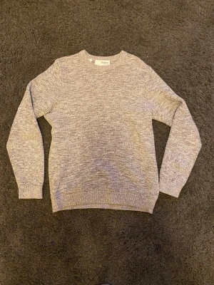 Grå stickad tröja från Selected Homme - Säljer en stilren gråmelerad stickad tröja från Selected Homme. Tröjan har rund halsringning, långa ärmar och ribbade muddar vid ärmslut och nederkant. Perfekt för lager på lager och enkel att matcha med olika outfits.