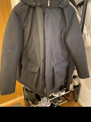 Svart parkas från BOSS med fickor - Snygg svart parkas från BOSS med flera stora fickor framtill, dragkedja och knappar. Jackan har hög krage och huva, perfekt för kyliga dagar. Klassisk och stilren design med diskret BOSS-logga på insidan och på armen.