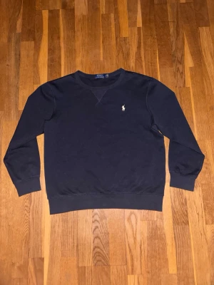 Mörkblå sweatshirt från Polo Ralph Lauren - Mörkblå sweatshirt från Polo Ralph Lauren med klassisk rund halsringning och broderad vit logga på bröstet. Tröjan har ribbade muddar vid ärmslut och nederkant. Perfekt för en avslappnad och stilren look.