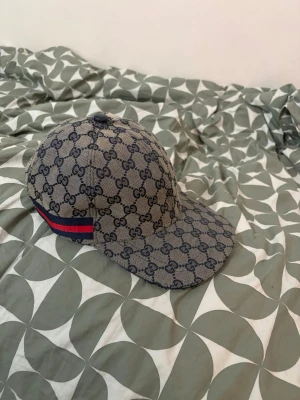  keps med GG-mönster - Säljer en snygg keps i beige och marinblått med klassiskt GG-monogram över hela kepsen. Kepsen har en böjd skärm och en röd-blå rand på sidan. Justerbar rem baktill och insida i mörkblått tyg. Perfekt för dig som gillar streetwear och exklusiva accessoarer.
