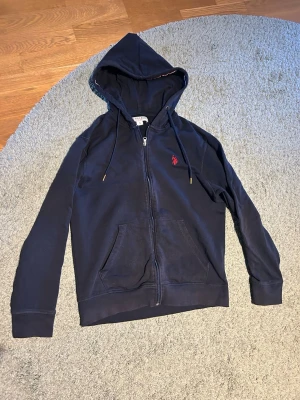 Marinblå hoodie från U.S. Polo Assn  - Snygg marinblå hoodie från U.S. Polo Assn med dragkedja, huva med dragsko och kängurufickor. Liten röd logga broderad på bröstet och diskret randigt foder i huvan. Perfekt för en chill och avslappnad stil. Pris kan diskuteras 😁