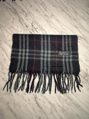 Halsduk från Burberrys  - Mörkgrå halsduk från Burberrys i 100% ull med klassiskt rutmönster i grått, svart och rött. Halsduken har fransar i båda ändar och en vävd logotypetikett. Perfekt accessoar för att lyfta din outfit.