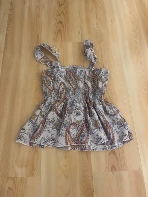 Mönstrad linnetopp med volang - Söt linnetopp med smockad överdel och volangig nederdel. Toppen har breda axelband och ett paisleyinspirerat mönster i beige, rött och gult. Perfekt för varma dagar och ger en bohemisk vibe.