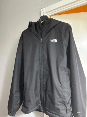 Svart regnjacka från The North Face - Snygg svart regnjacka från The North Face med huva och dragkedja framtill. Jackan har logga på bröstet och är tillverkad i ett vattenavvisande material. Perfekt för regniga dagar och enkel att matcha med olika outfits.