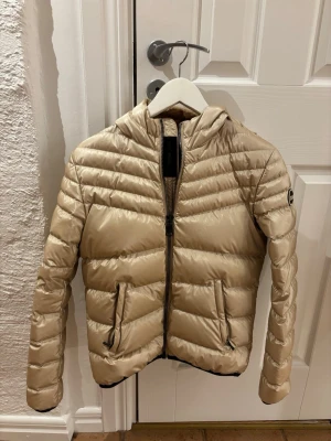 Beige dunjacka med huva - Snygg beige dunjacka från Colmar  med huva och dragkedja framtill. Jackan har två fickor med dragkedja och quiltad design. 