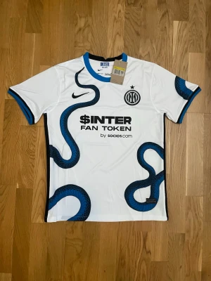 Inter Milan bortatröja Nike 21/22 - Säljer en vit Inter Milan bortatröja från Nike säsongen 21/22 med blå och svart orm-mönster. Tröjan har korta ärmar, svart och blå kant vid ärmslut och tryckta loggor från Inter, Nike, $INTER och Lenovo. Tillverkad i lätt och ventilerande material.