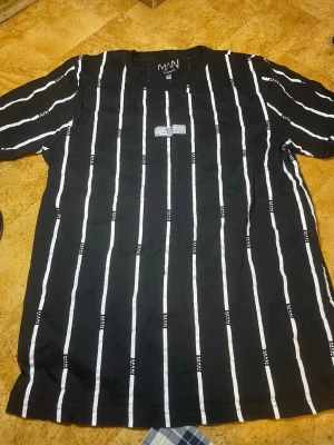 Svart randig t-shirt från MAN - Snygg svart t-shirt från MAN med vita vertikala ränder och ett litet tygmärke på bröstet. T-shirten har rund hals och korta ärmar. Perfekt för en trendig och avslappnad stil.