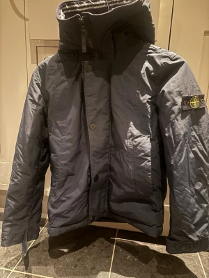 Mörkblå parkas från Stone Island - Snygg mörkblå parkas från Stone Island med ikonisk patch på ärmen. Jackan har hög krage, dragkedja och knappar framtill samt två stora fickor. Materialet är slitstarkt och jackan har en modern, rak passform med justerbar huva.