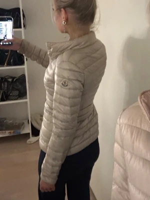 Beige dunjacka från Moncler - Snygg beige dunjacka från Moncler med quiltad design och hög krage. Jackan har tryckknappar framtill, två fickor och den klassiska Moncler-loggan på ärmen. Perfekt för kalla vinterdagar och ger en stilren look. Jag köpte denna på plick för 290€!