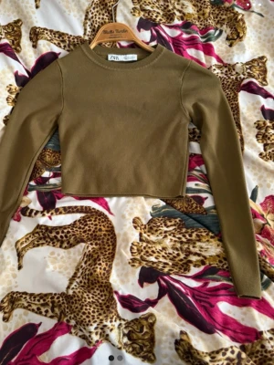 Olivgrön croppad topp från Zara - Trendig olivgrön croppad topp från Zara med lång ärm och rund halsringning. Enkel och stilren design som passar perfekt till höga jeans eller kjol. Tillverkad i mjukt material för en bekväm känsla. S men känns som XS