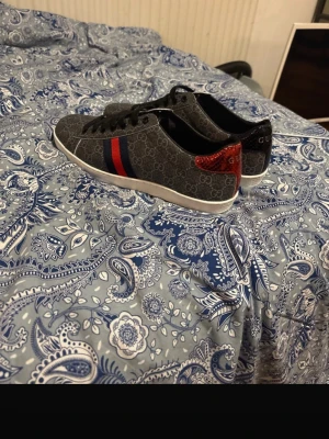 Gucci sneakers med GG-mönster - Snygga sneakers från Gucci i mörkgrått tyg med klassiskt GG-mönster. Skorna har svarta snören, vit sula och de ikoniska röd-blå ränderna på sidan. Baktill finns detaljer i rött och blått ormskinnsmönster samt Gucci-logga. Perfekta för dig som gillar lyxig streetstyle.