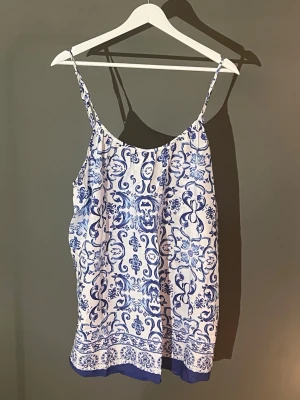 Vit och blå mönstrad linnetopp - Säljer en snygg linnetopp med tunna axelband och ett blått, dekorativt mönster på vit botten. Toppen har en lös passform och en blå bård längst ner. Perfekt för varma dagar och enkel att styla till jeans eller kjol🩵💙