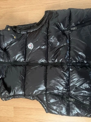 Svart dunväst från Moncler - Snygg svart dunväst från Moncler med glansig finish och ikonisk logga på bröstet. Västen har dragkedja framtill, två sidofickor med dragkedja och en hög krage. Perfekt för lager-på-lager och streetstyle.