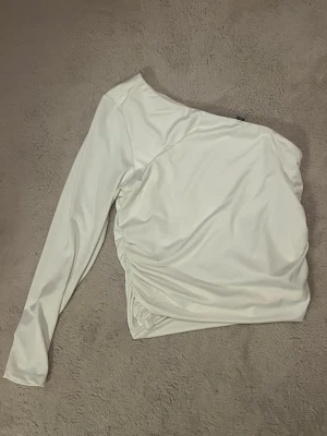Vit asymmetrisk topp med rynk - Trendig vit topp med en axel och lång ärm. Toppen har snygga rynkdetaljer på sidan som ger en cool och modern look. Perfekt för dig som vill sticka ut lite extra med en unik form🤍