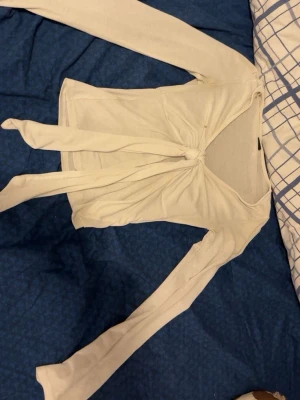 Vit omlottopp med knyt framtill - Trendig vit omlottopp med långa ärmar och knyt framtill som ger en snygg v-ringad look. Toppen är croppad och har en mjuk, stretchig känsla. Perfekt att styla med jeans eller kjol för en cool och avslappnad vibe.