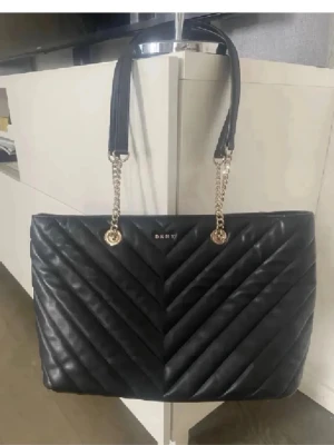 Svart quiltad handväska från DKNY - Stilren svart handväska från DKNY med quiltat mönster och guldfärgade kedjedetaljer på axelbanden. Väskan har en rektangulär form och är tillverkad i läder. Perfekt för dig som vill ha en trendig och elegant accessoar.