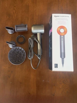 Dyson Supersonic hårfön med tillbehör - Säljer en Dyson Supersonic hårfön i silver och orange med flera stylingmunstycken. Hårfönen har en modern, rund design och levereras med originalkartong. Perfekt för dig som vill ha snabb och skonsam hårtorkning med avancerad teknik.