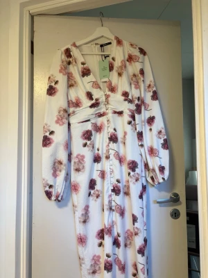 Vit blommig långklänning med v-ringning - Långklänning i vit bas med blommigt mönster i rosa och vinröda toner. Klänningen har v-ringning, långa puffärmar och knappar framtill. Tillverkad i ett lätt, strukturerat tyg som ger en elegant känsla. Perfekt för sommarens alla tillfällen.