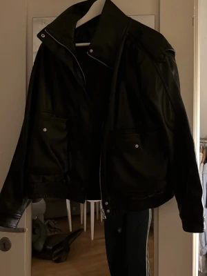 Svart skinnjacka från Gina Tricot - Snygg svart skinnjacka från Gina Tricot med två stora fickor framtill och silverfärgade knappar. Jackan har en rak passform, hög krage och dragkedja. Perfekt för dig som vill ha en klassisk och stilren look.