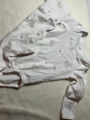 Vit pyjamas med kaninmönster H&M - Söt vit långärmad pyjamas från H&M med små kaniner och blommor tryckta över hela plagget. Tillverkad i mjuk bomull, perfekt för mysiga kvällar och nätter. Klassisk rund hals och tryckknappar i grenen för enkel påklädning.