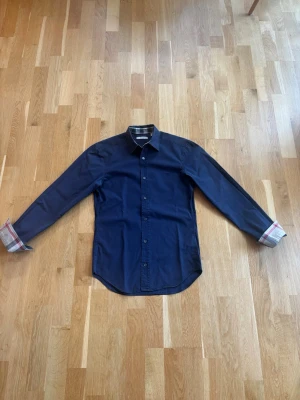 Mörkblå skjorta från Burberry - En stilren mörkblå skjorta från Burberry med de klassiska rutmönstret i kragen och på insidan av ärmarna. Den är i väldigt fint skick utan några skador. De ända är att den är lite blekt men det är inget man tänker på. Hör gärna av er vid minsta fundering.