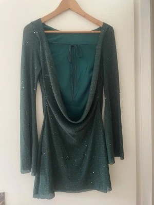 Grön glittrig kortklänning med öppen rygg - Snygg grön kortklänning med glittriga detaljer och djup öppen rygg med snörning. Klänningen har lång ärm och ett lätt utställt fall. Perfekt för dig som vill sticka ut med en unik look. Storlek S.